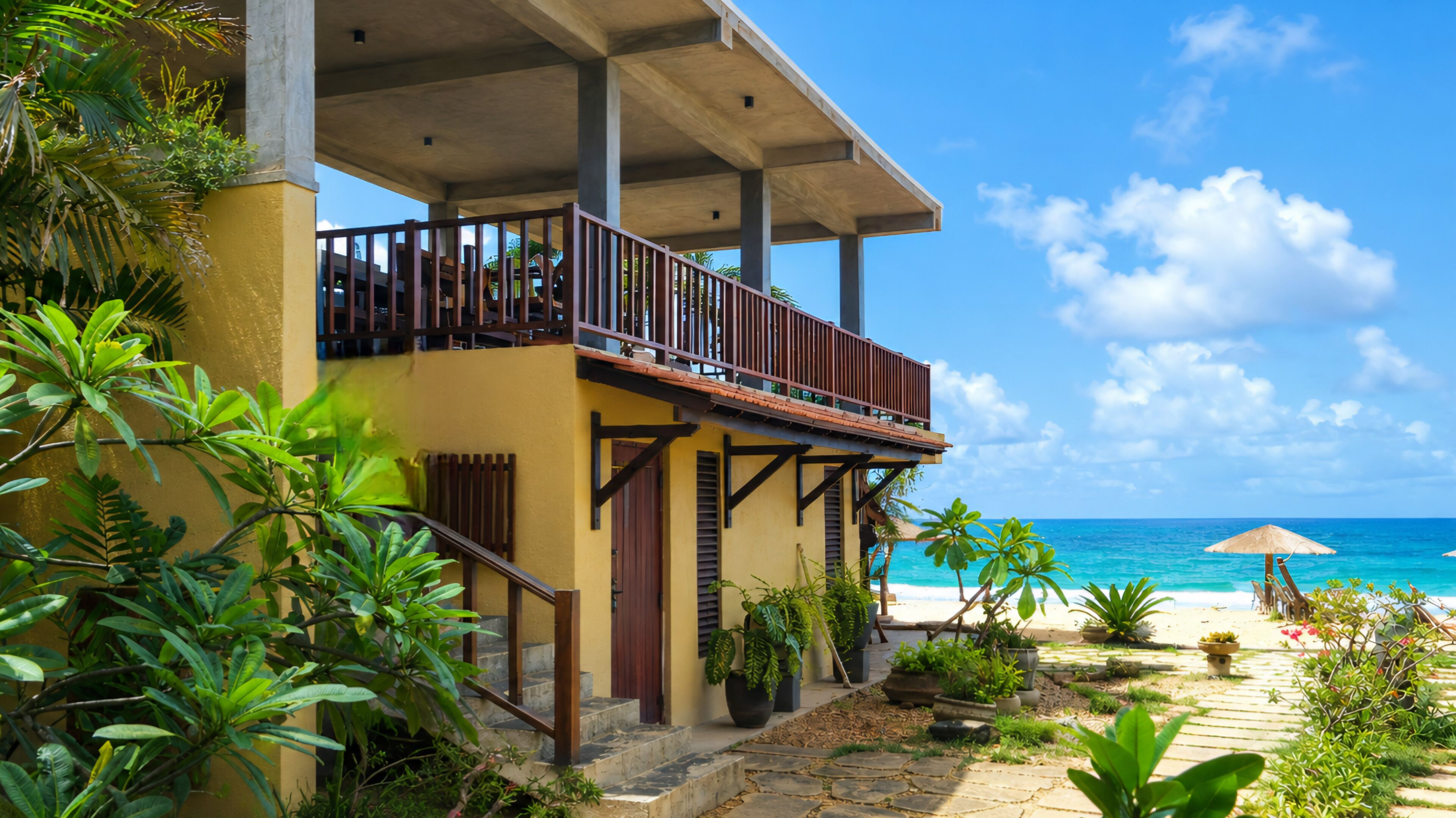 Un immeuble de deux étages doté d’un balcon donnant sur une plage, entouré de plantes tropicales et surplombé d’un ciel bleu azur.