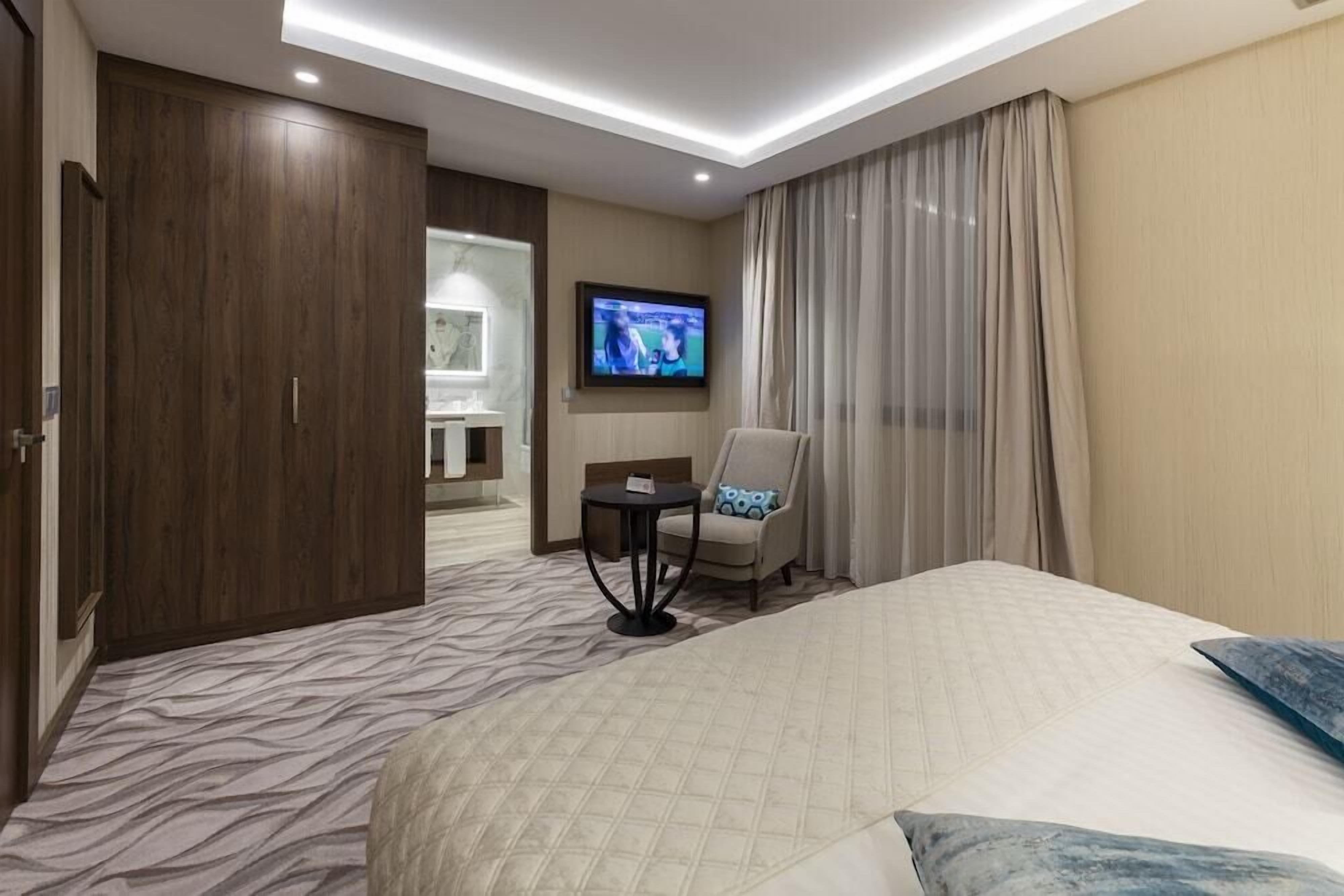 A hotel room with a bed, a chair, a TV, and a bathroom visible through an open door.