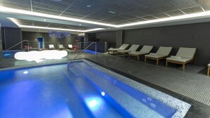 An indoor swimming pool with a tiled floor, a changing room with a shower, and lounge chairs along the poolside. - AZ Grand Oran Hotel (Es Senia)