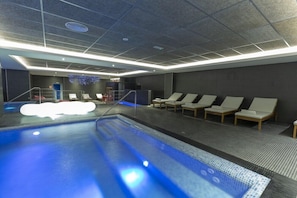 Een binnenzwembad met betegelde vloer, een kleedruimte met douche en loungebedden langs het zwembad.