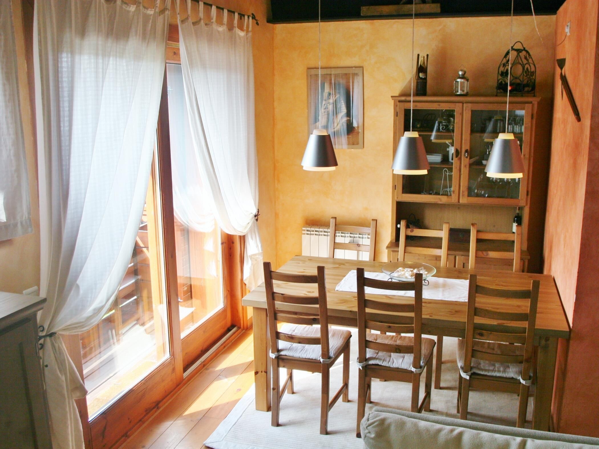 Un comedor con mesa y sillas de madera, un aparador con puertas de cristal y una ventana con cortinas.