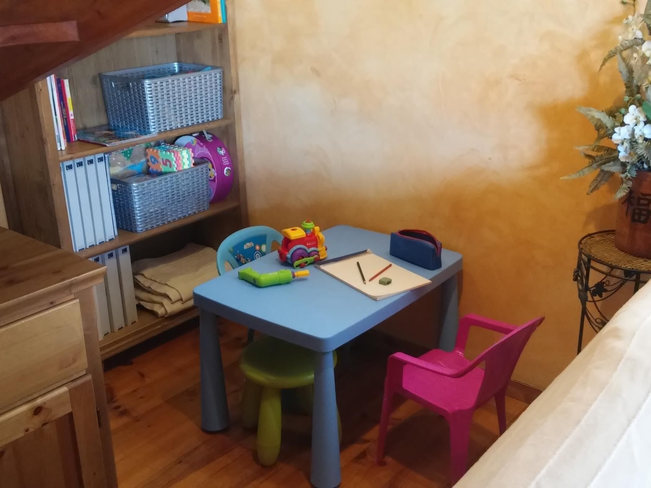 Una habitación pequeña con una mesa azul, una silla rosa y un estante de madera con cajas de almacenamiento.