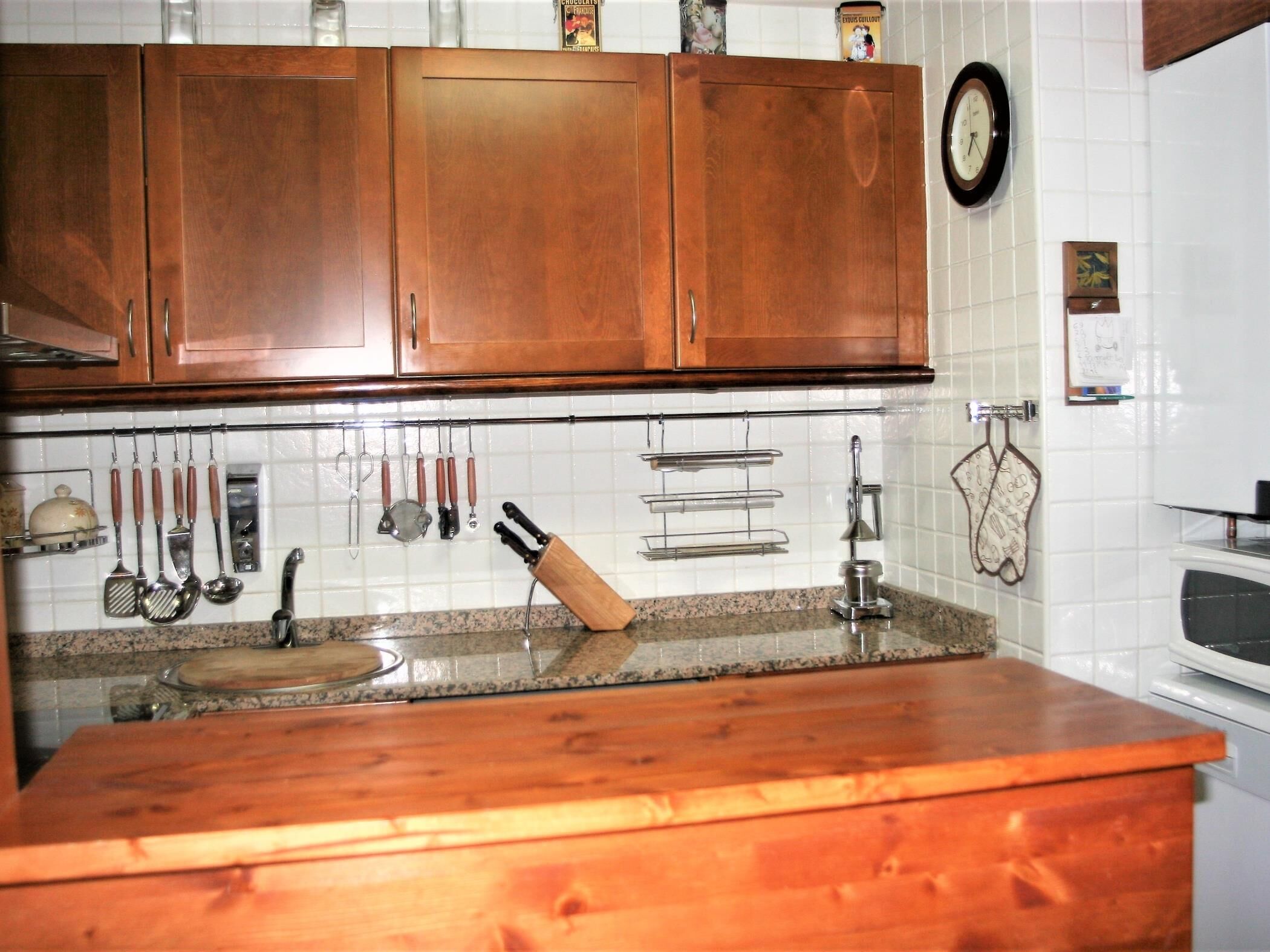 Una cocina con armarios de madera, encimera de granito y diversos utensilios colgando sobre el fregadero.