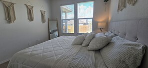 A bedroom with a large bed, a window with blinds, a bedside lamp, and macramé wall hangings. - Family-Friendly New Townhome in lovely Wayside community (Uhland)