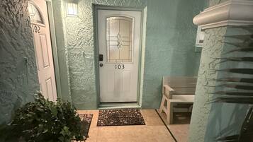 Un pasillo con una puerta identificada con el número 103, un banco, una planta en maceta y una pared con textura.