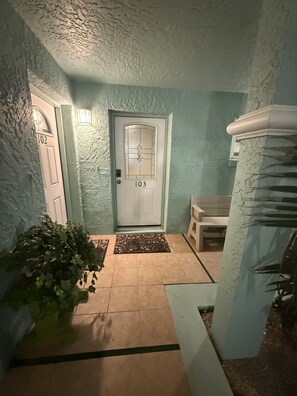 A hallway with a door labeled 103, a bench, a potted plant, and a textured wall. - Daytona Riverfront Haven Retreat on the Halifax (Port Orange)