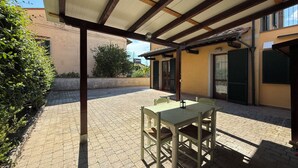 Een overdekte patio buiten met een tafel en stoelen, een betegelde vloer, en een gebouw met een borduurwerk in de achtergrond.
