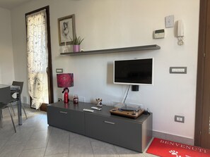 A modern living room with a flat-screen TV, a console table, a red lamp, and a 'BENVENUTI' welcome mat.