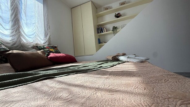Een slaapkamer met een bed, een kledingkast, een plank met boeken en een plant, en een raam met dunne gordijnen.