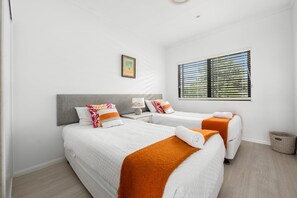 Habitación de hotel con dos camas, una ventana con persianas, un cuadro enmarcado en la pared y una cesta pequeña en el suelo.