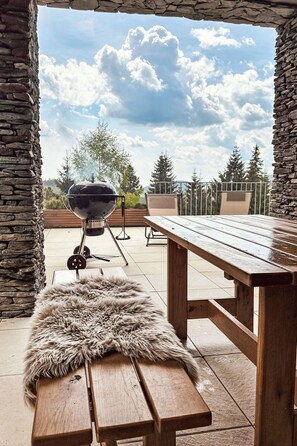 An outdoor patio with a stone wall, a wooden table, a fur rug, and a barbecue grill. - Rebelka by Interhome (Horni Misecky)