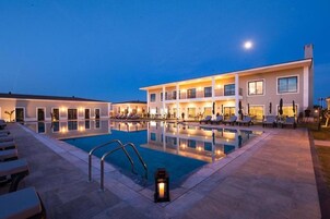 Un hotel con piscina, sillones de descanso y fachada iluminada al anochecer.