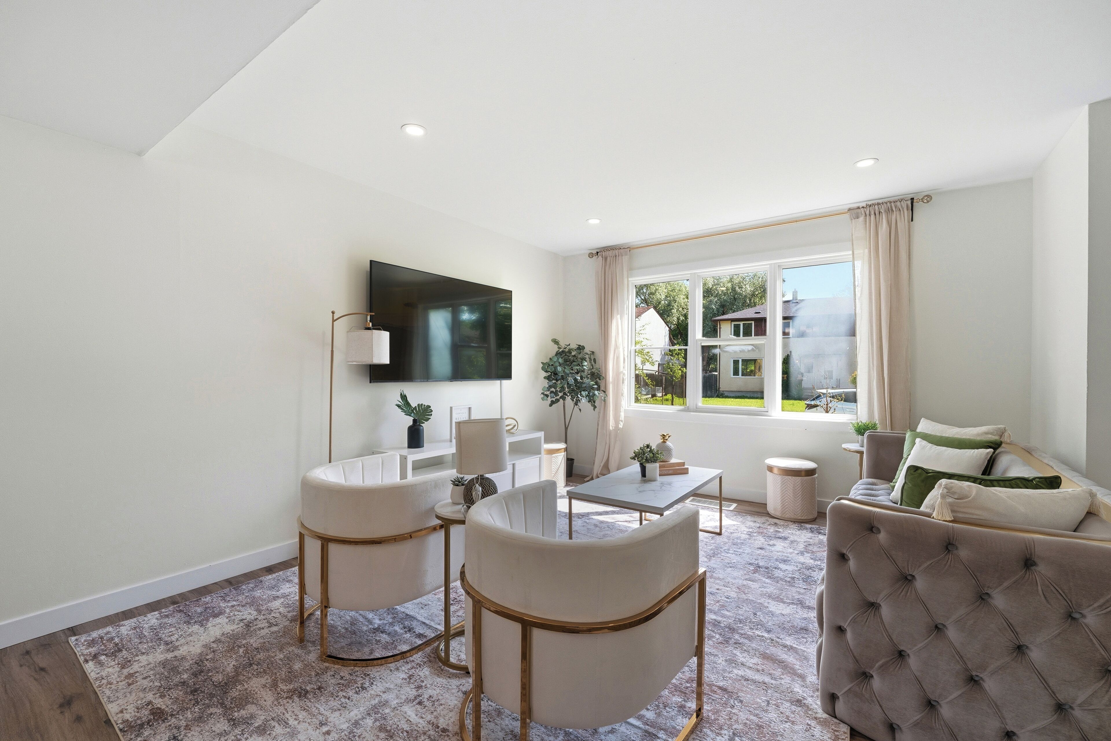 A modern living room with a flat-screen TV mounted on the wall, a round coffee table, two armchairs, a sofa, and a large window with curtains.