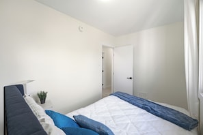 Un dormitorio con cama, mesita de noche, planta en maceta y puerta que da a otra habitación.