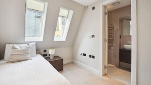 A bedroom with a bed, bedside table, lamp, and two windows. - Buckingham Palace Residences by Aeria Apartments (London)