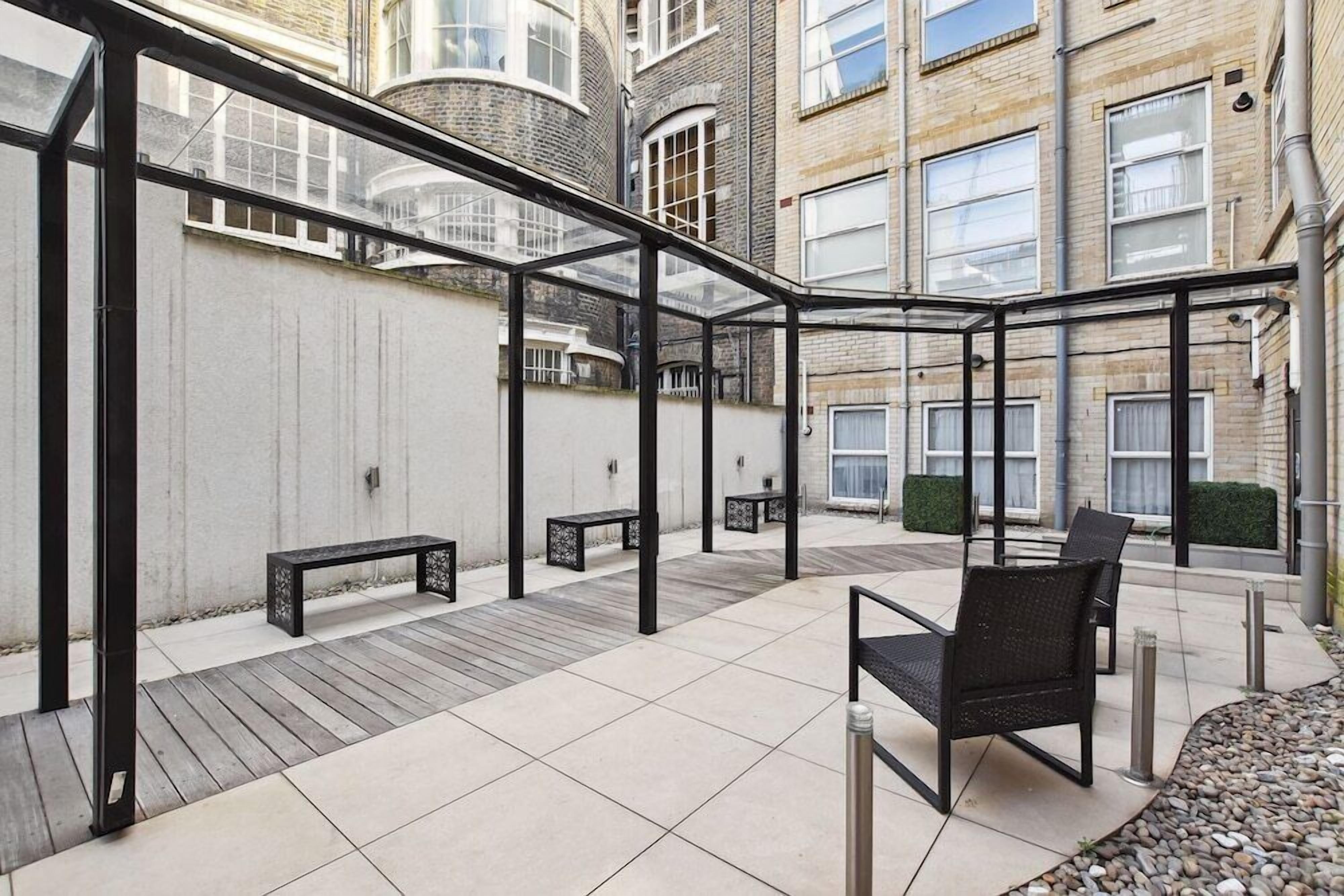 A covered outdoor seating area with benches and chairs, a wooden deck, and a view of a brick building.