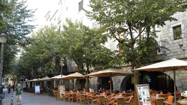 Ein Straßencafé mit Tischen und Stühlen, umgeben von Bäumen und Gebäuden.