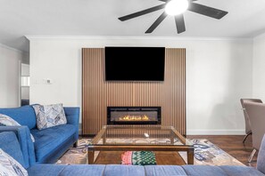 A living room with a fireplace, a blue sofa, a glass coffee table, and a ceiling fan. - Euless 4BR Home - Mini Golf, Hot Tub & Arcade - Near DFW Airport (Euless)