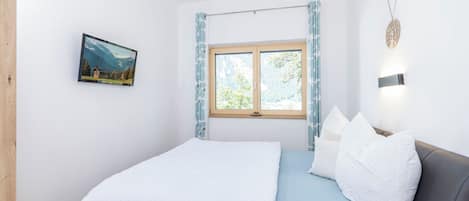 A bedroom with a wooden bed, a window with curtains, a wall-mounted picture, and a wall light.