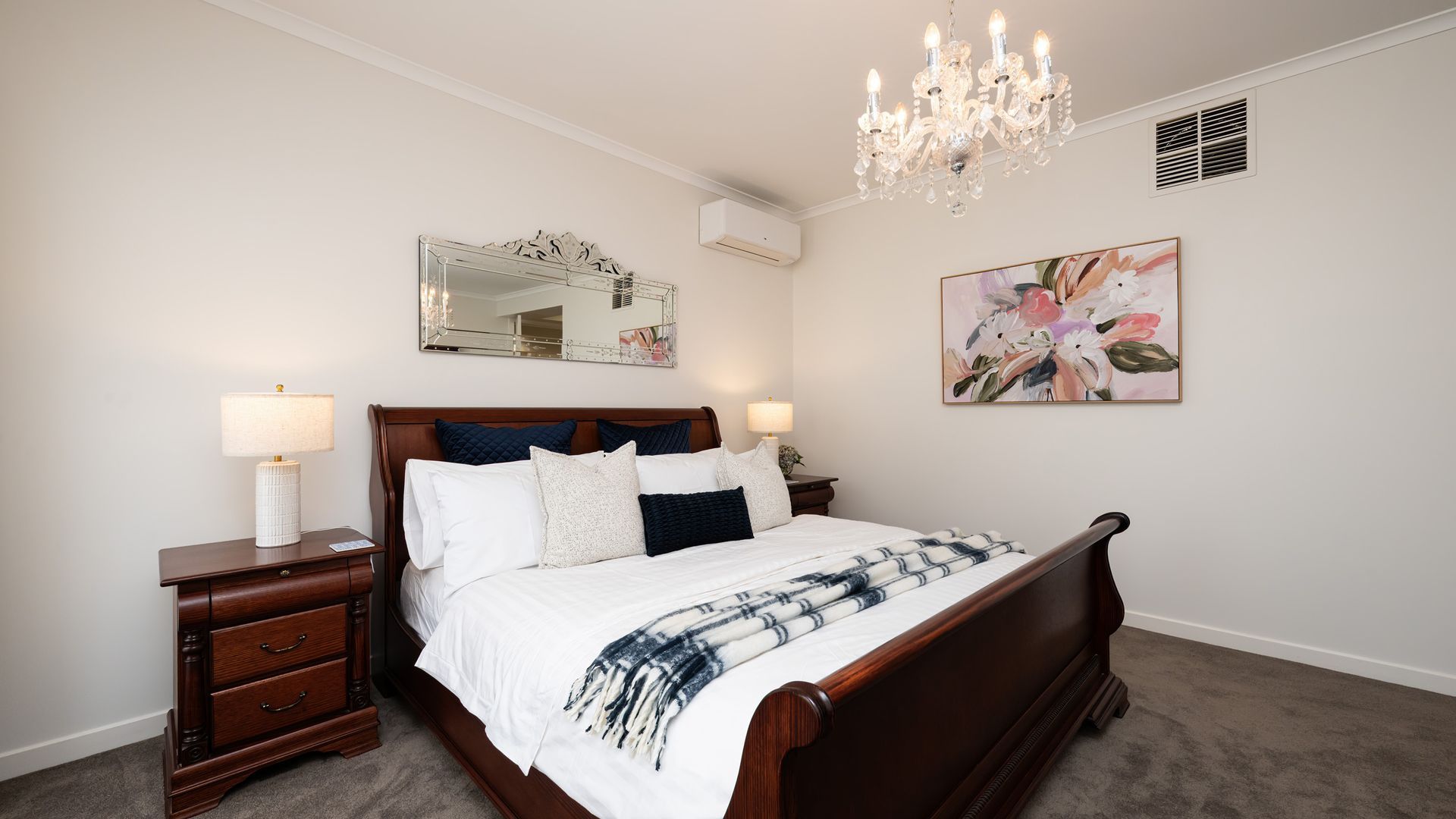 A bedroom with a wooden bed, two bedside tables with lamps, a chandelier, a wall-mounted air conditioner, and a floral painting.