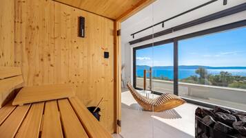 Ein Holzsaunaraum mit einer Bank und einer hängenden Holzliege, große Fenster mit Blick auf das Meer und die Berge.