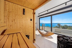 Un sauna de madera con banco y asiento colgante, ventanas amplias con vista al mar y las montañas.