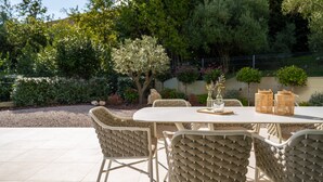 Eine Terrasse im Freien mit einem runden Tisch und Korbstühlen, umgeben von Grünpflanzen und einem Zaun.