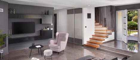 A modern living room with a flat-screen TV, a sofa, a chair, a coffee table, and a staircase leading to an outdoor area.