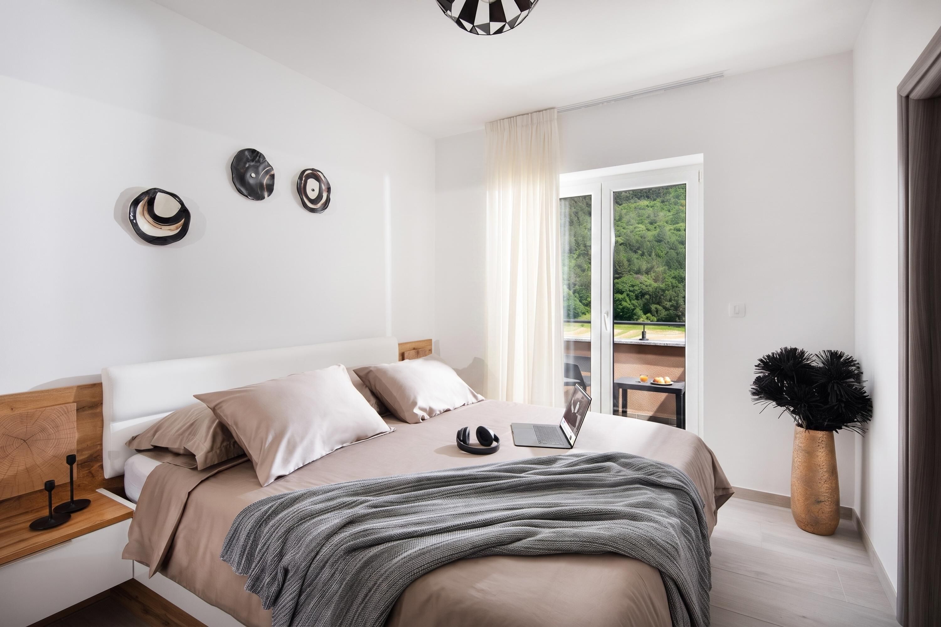 A modern bedroom with a bed, a laptop, and a view of a balcony with greenery.