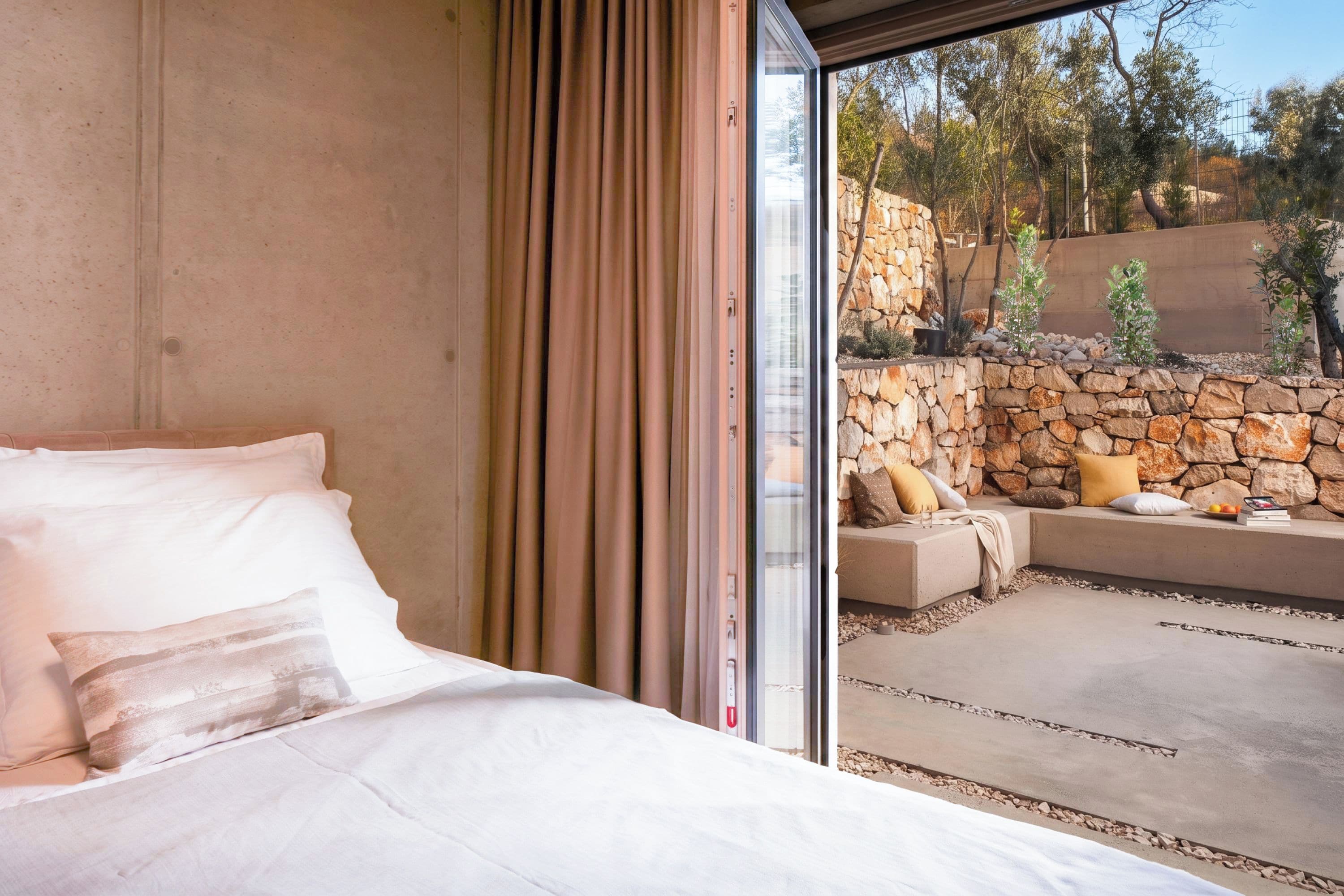 Un dormitorio con una cama grande, una puerta corrediza de vidrio que da a un espacio exterior con área de descanso, una pared de piedra y plantas.