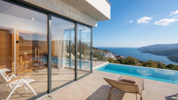 A modern outdoor pool area with glass walls, a view of the sea, and a clear sky. - Villa Saturea With Pool & Sauna Near Rabac (Labin)