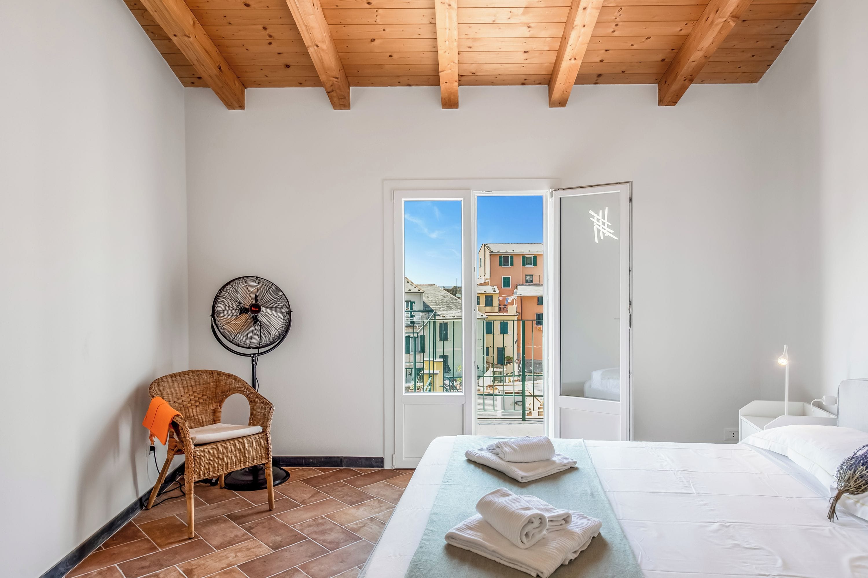 Una camera da letto con un letto, una sedia, un ventilatore e vista sugli edifici attraverso una porta.