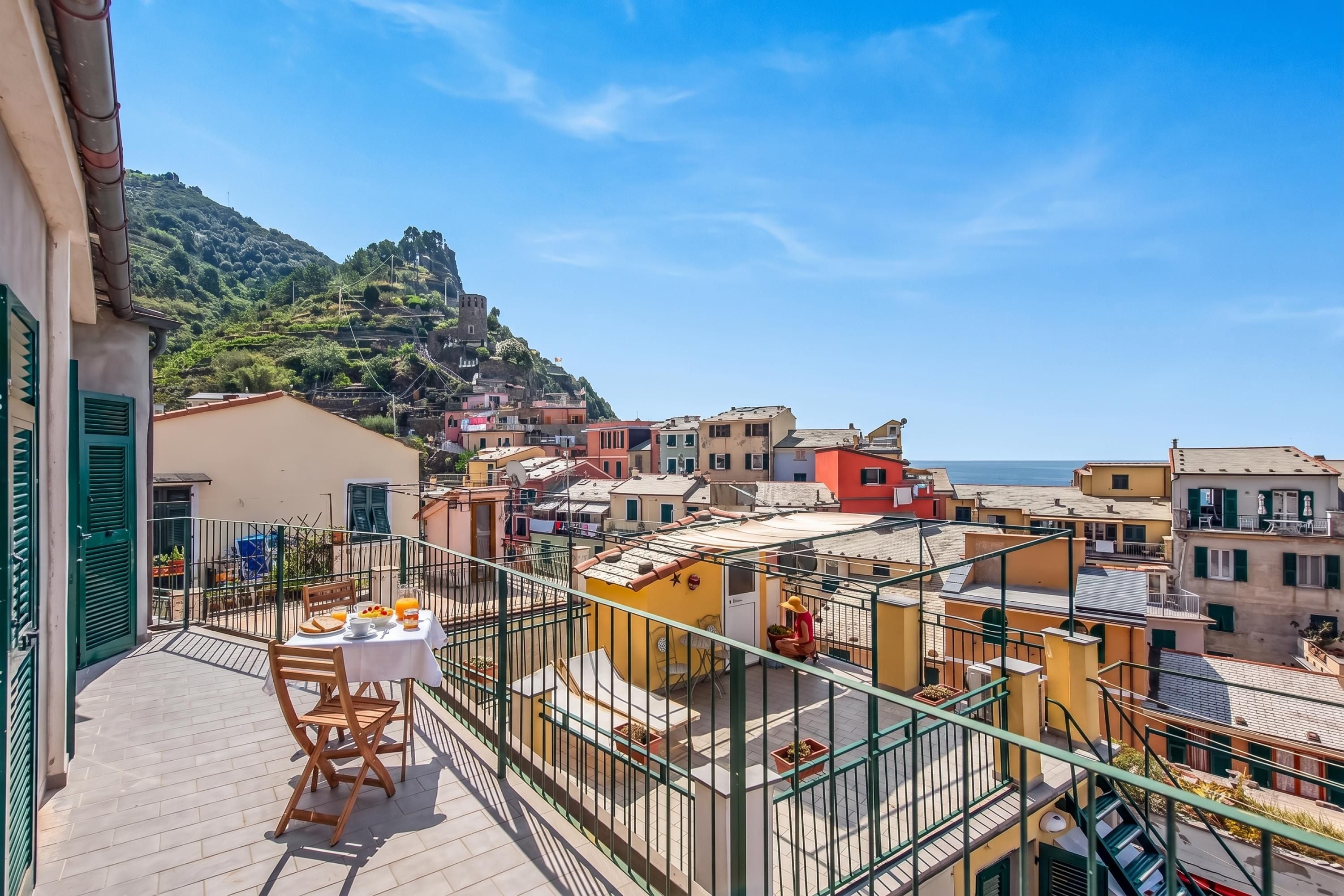 A balcony with a table set for breakfast, overlooking a coastal town with colorful buildings and a hill in the background.
