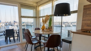 A modern dining area with a view of a marina, featuring a round table, four chairs, a floor lamp, and a small kitchen area. - Enjoy in the harbor at Veerse Meer. (Wolphaartsdijk)