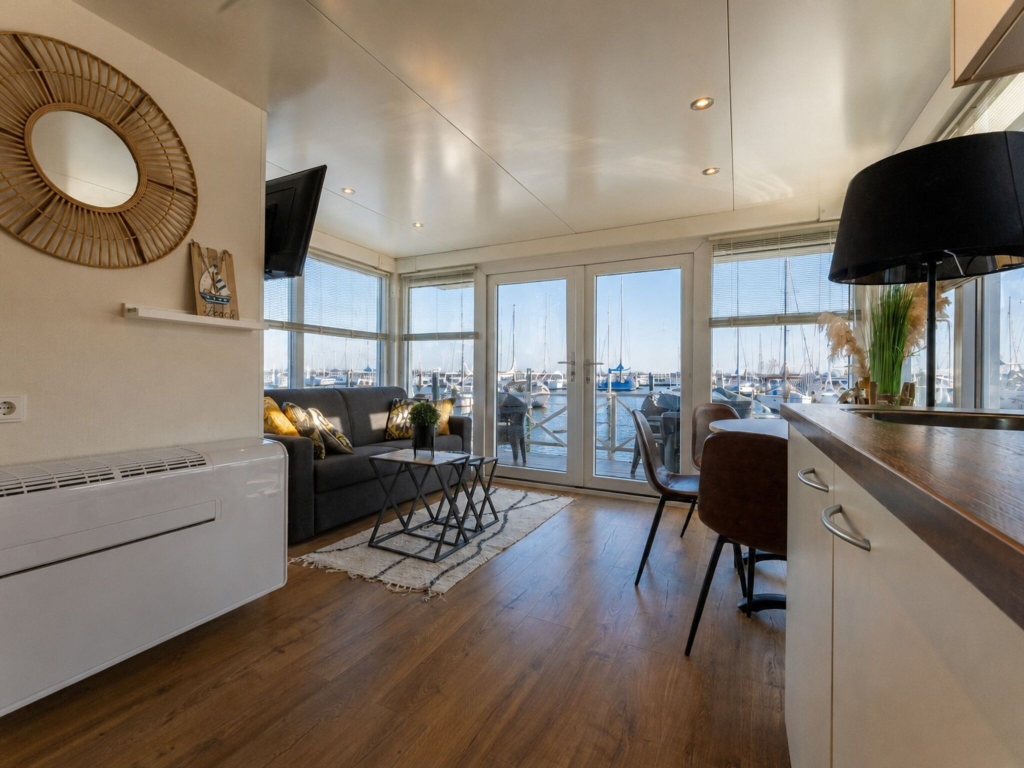 Un espacio habitable moderno con sofá, mesa de comedor, sillas, ventanas amplias con vista al puerto deportivo y una cocina con estufa.