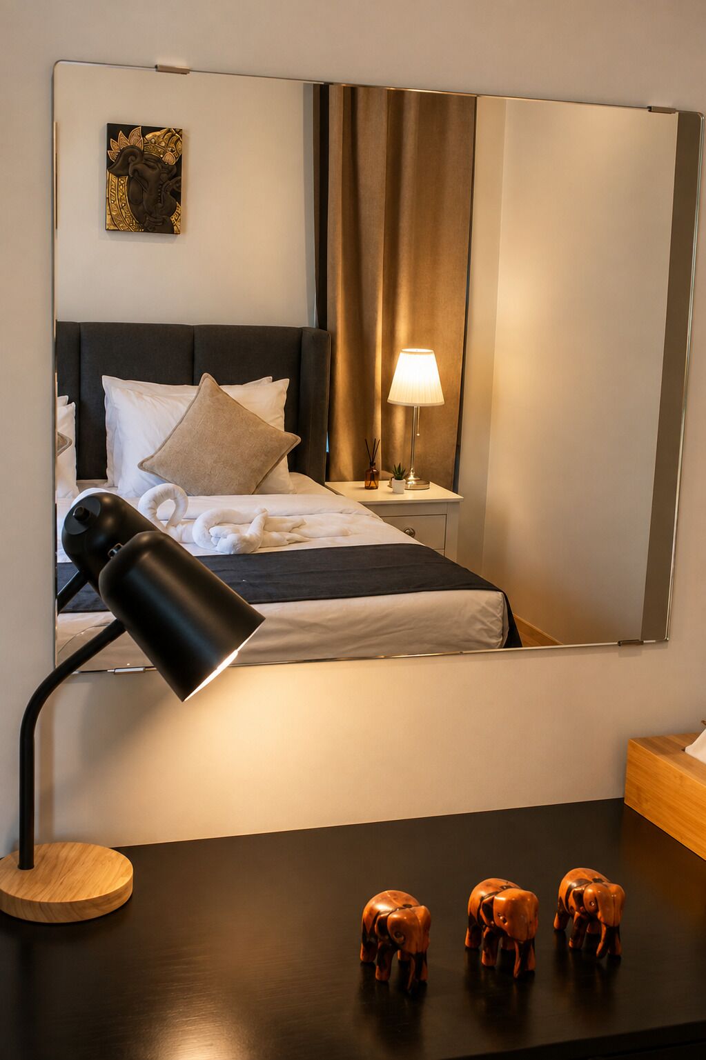 A hotel room with a bed, a bedside lamp, a nightstand, a mirror, and decorative elephant figurines.