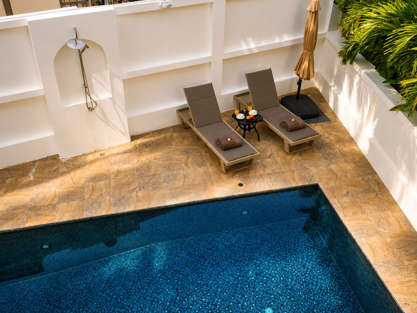 A swimming pool with lounge chairs and a table with drinks, surrounded by a white wall and plants.