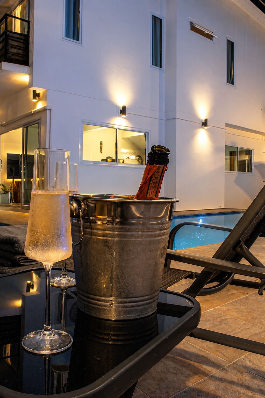 Zona junto a la piscina con una mesa de bar, un vaso de bebida transparente, un cubo metálico con una botella y una piscina al fondo.