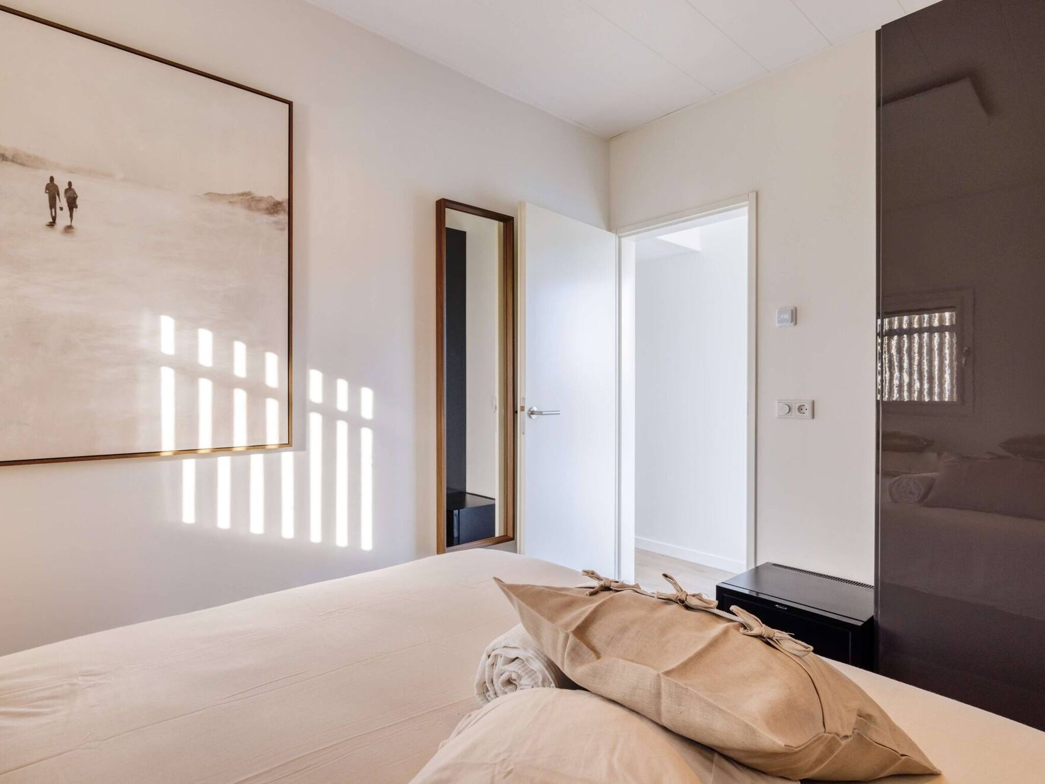 Ein Schlafzimmer mit einem Bett, einem Nachttisch, einer Lampe, einer Tür und einem an der Wand befestigten Kunstwerk.