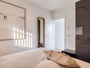 A bedroom with a bed, a bedside table, a lamp, a door, and a wall-mounted artwork.