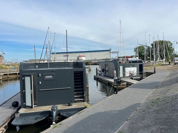 Un port de plaisance avec des bateaux amarrés, un bâtiment portant l’inscription « VEKA » à l’arrière-plan, et une promenade piétonne le long de la berge.