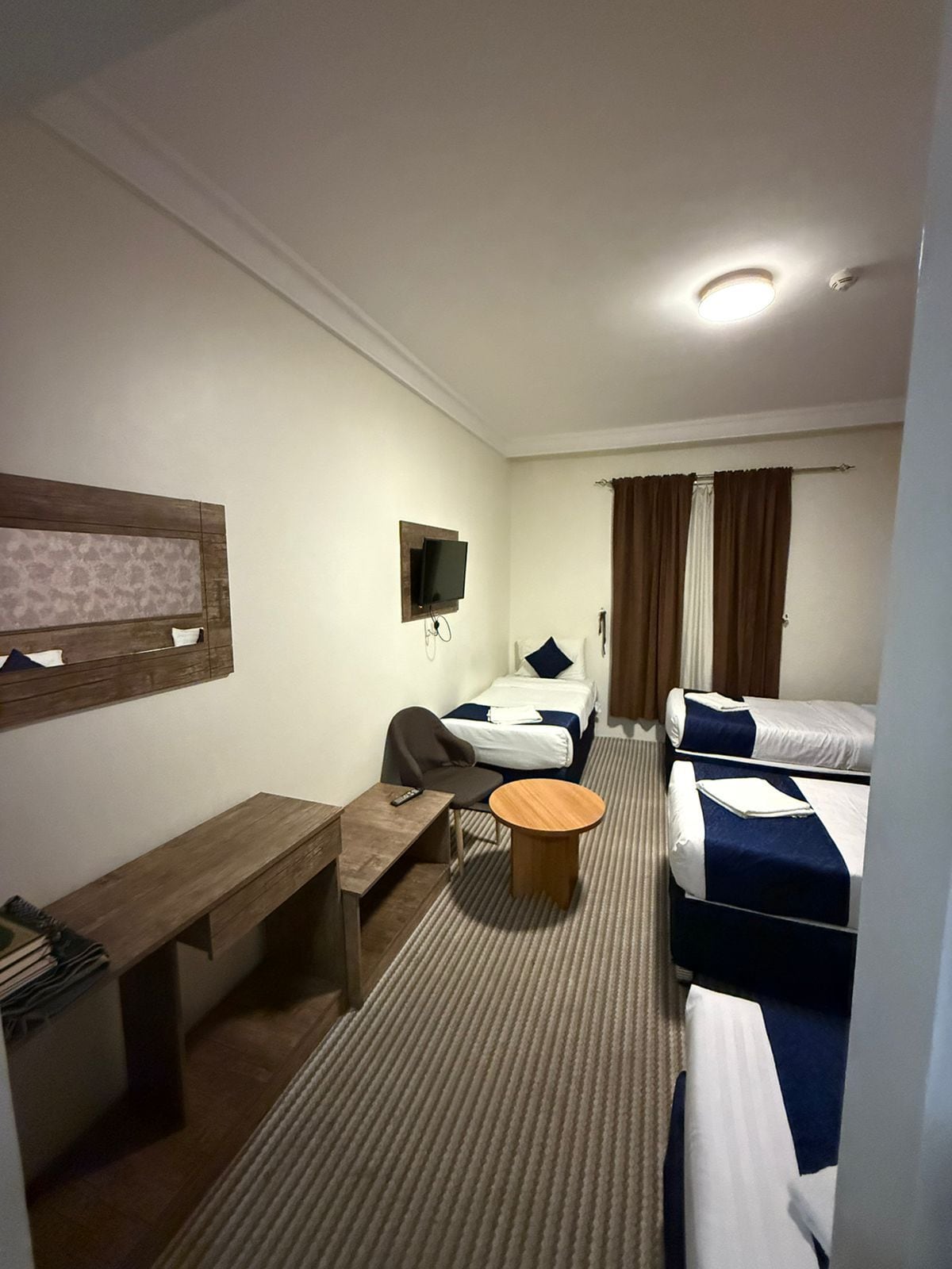 A hotel room with a single bed, a double bed, a desk with a chair, a small round table, and a television mounted on the wall.