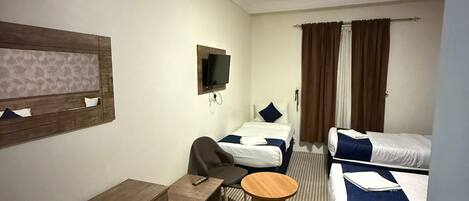 A hotel room with a single bed, a double bed, a desk with a chair, a small round table, and a television mounted on the wall.