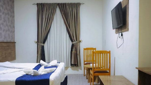 A hotel room with a bed, a wooden chair, a small table, a television mounted on the wall, and a window with curtains.