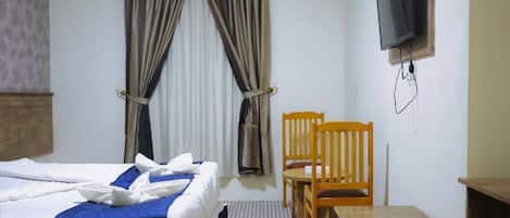 Habitación de hotel con cama, una silla de madera, una mesita, un televisor montado en la pared y una ventana con cortinas.