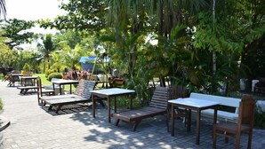 Een terras buiten met houten meubels en tafels, omringd door weelderige beplanting en palmbomen.