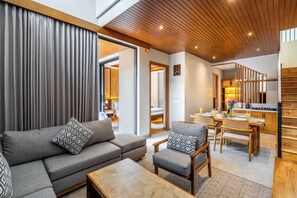 A modern living room with a sofa, armchair, and coffee table, a dining area with a table and chairs, and a kitchen with wooden cabinets and a stove. - Kanasu Villa by Exotic Bali Properties (Canggu)