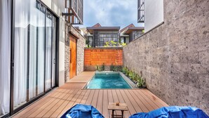 A modern outdoor pool area with a wooden deck, a small table, and two blue-covered lounge chairs. - Kanasu Villa by Exotic Bali Properties (Canggu)