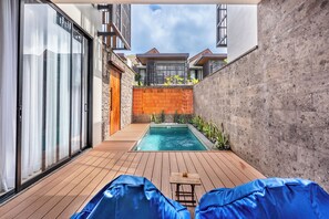 A modern outdoor pool area with a wooden deck, a small table, and two blue-covered lounge chairs. - Kanasu Villa by Exotic Bali Properties (Canggu)