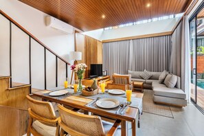 Ein modernes Wohnzimmer mit Essbereich, ausgestattet mit Holzbalkendecke, Flachbildfernseher, Sofa und Blick ins Freie.